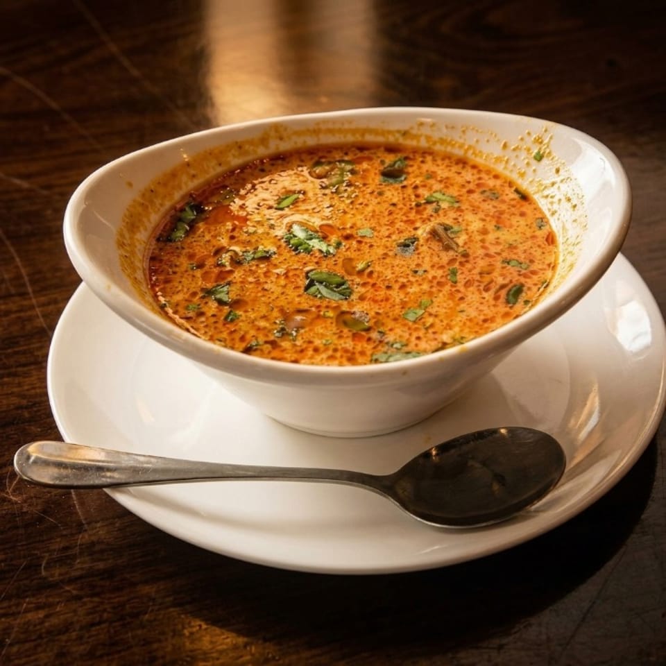A white bowl of creamy orange soup with green herbs sits on a white plate with a spoon.